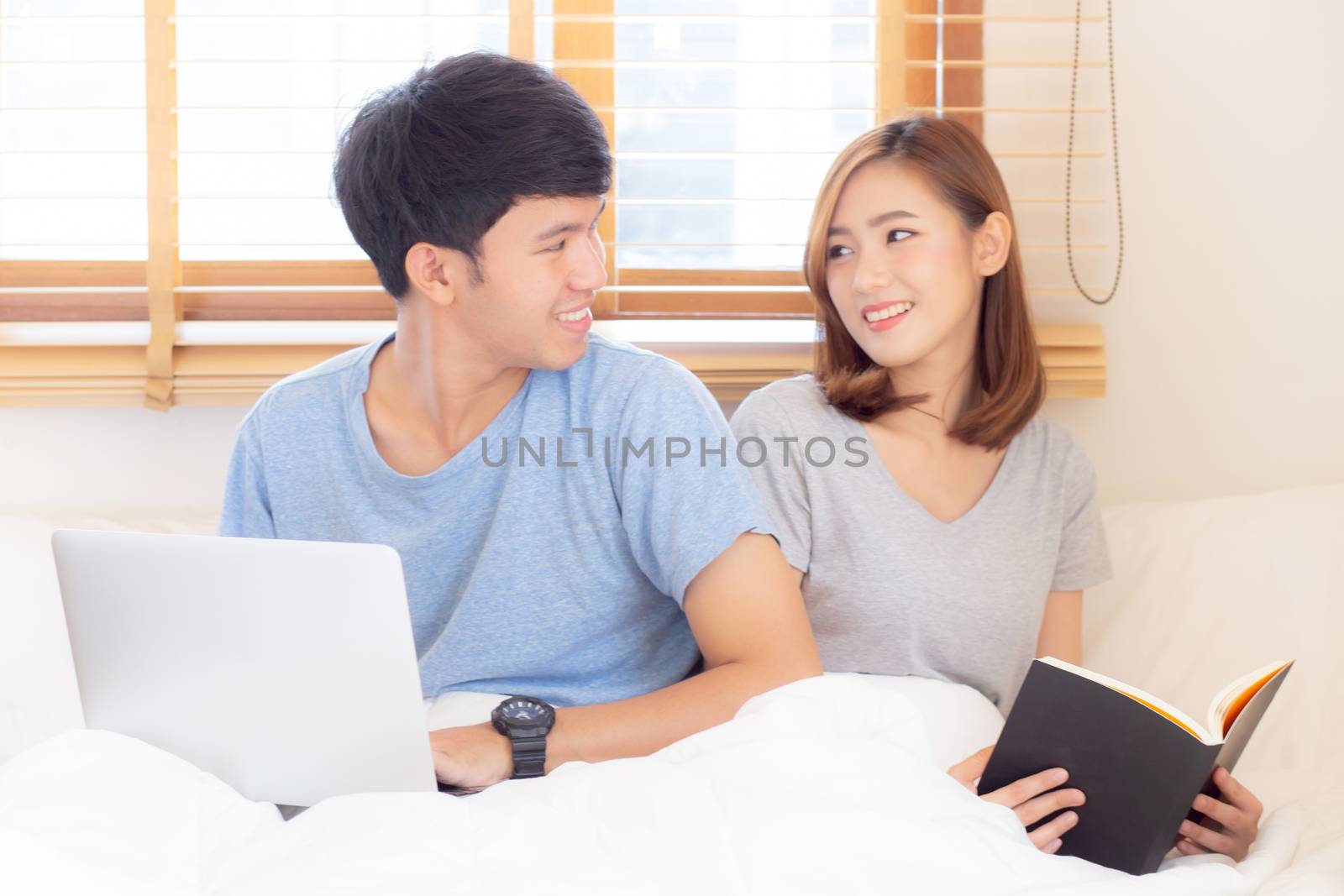 Asian young couple with man using laptop computer and searching internet and woman reading notebook on bed at bedroom with happy and relax, communication and business concept.