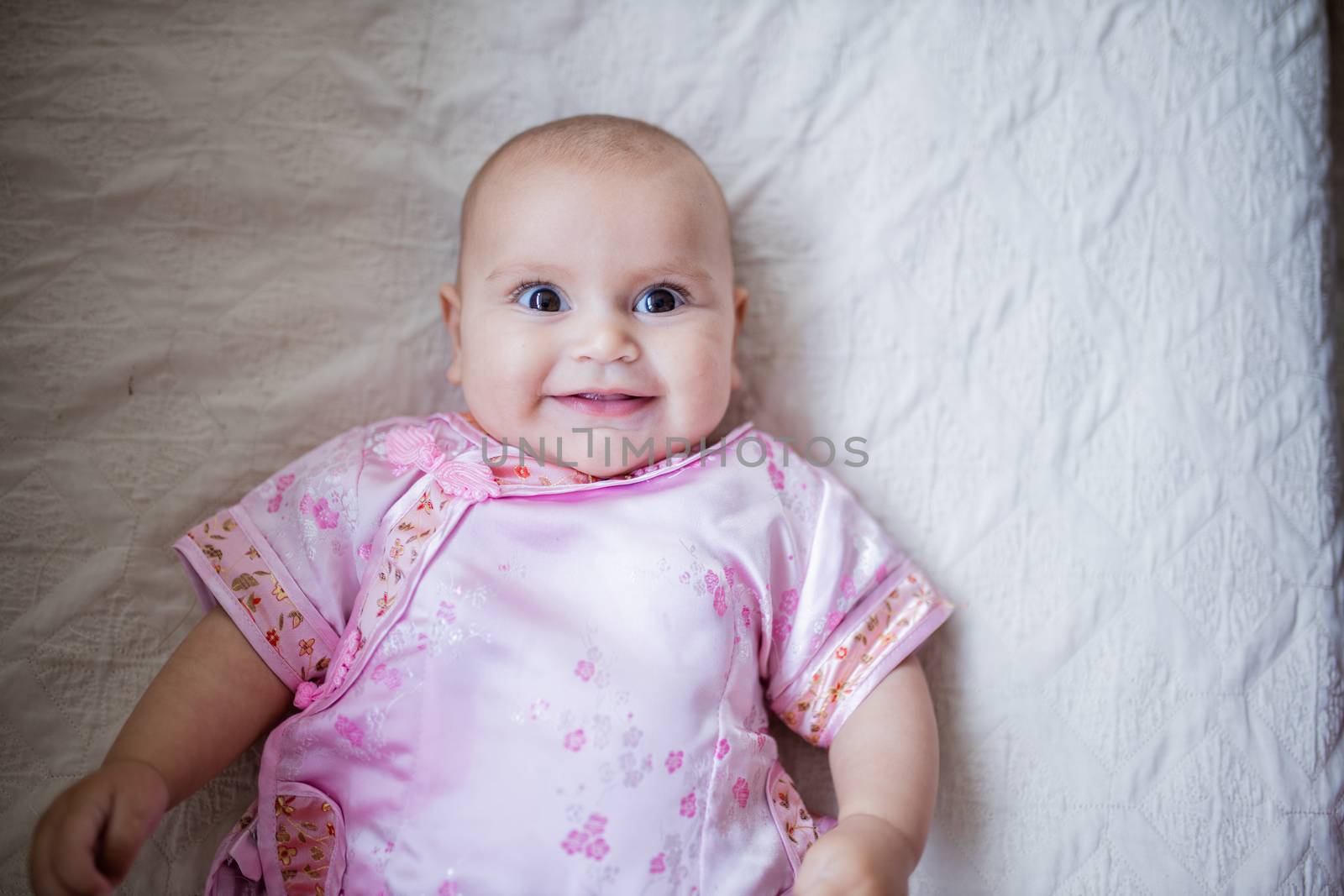 Happy baby girl in Asian pink attire lying down on a bed by Kanelbulle