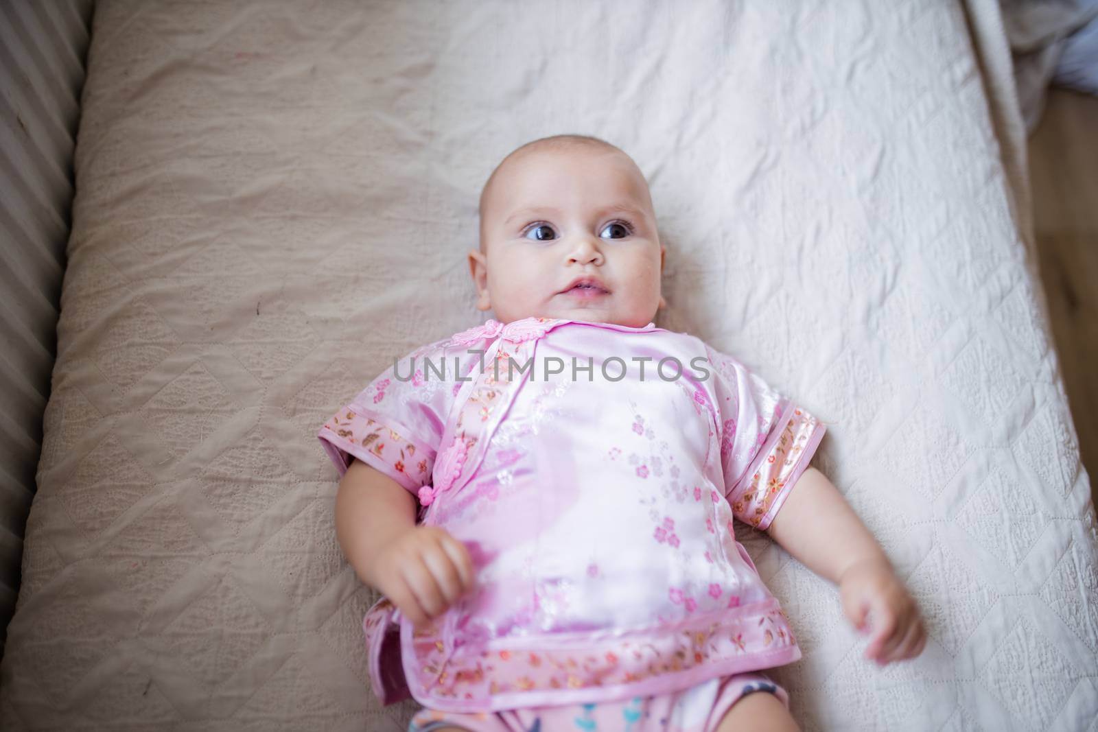 Curious adorable baby girl from above in Asian pink attire lying down on white bed. Portrait of cute female baby resting on bed. Happy babies on cradles and beds