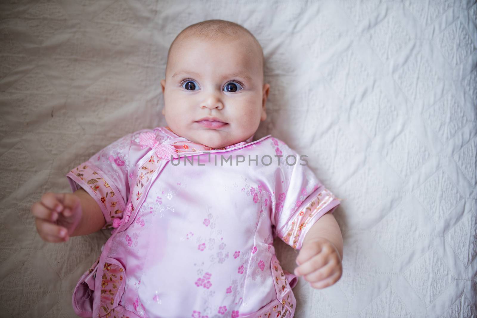 Curious and adorable baby girl in Asian pink attire lying down on a bed by Kanelbulle