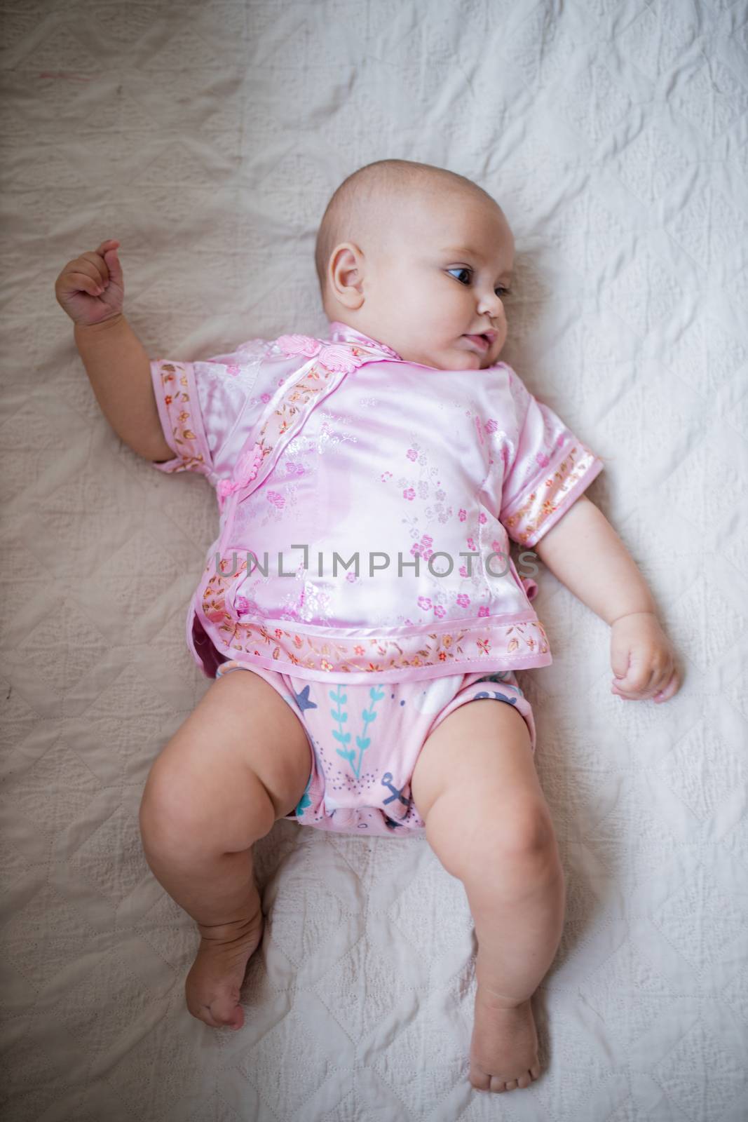 Distracted adorable baby girl from above in Asian pink attire lying down on white bed. Portrait of cute female baby resting on bed. Happy babies on cradles and beds