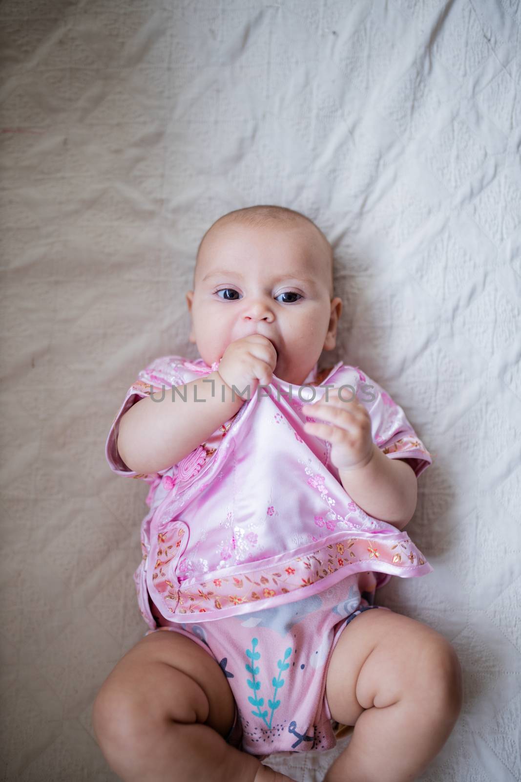 Adorable baby girl from above biting her Asian pink attire and lying down on white bed. Portrait of joyful female baby resting on bed. Happy babies on cradles and beds