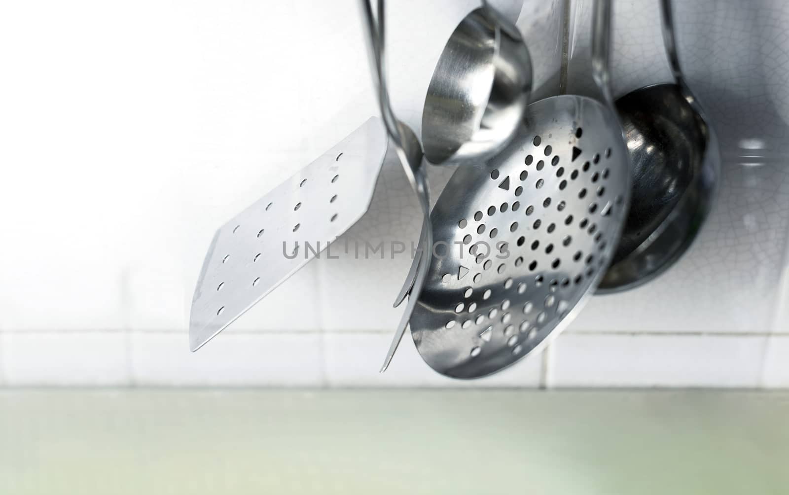 A skimmer, two forks and two metal ladles hanging on the tiled kitchen wall. Metal kitchen utensils