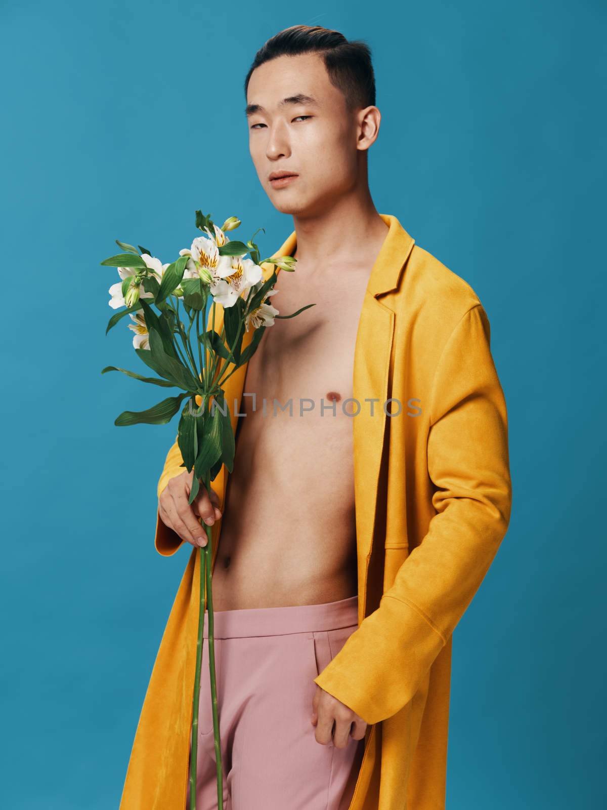 Asian man with bouquet of flowers on blue background and unbuttoned coat pink pants cropped view. High quality photo