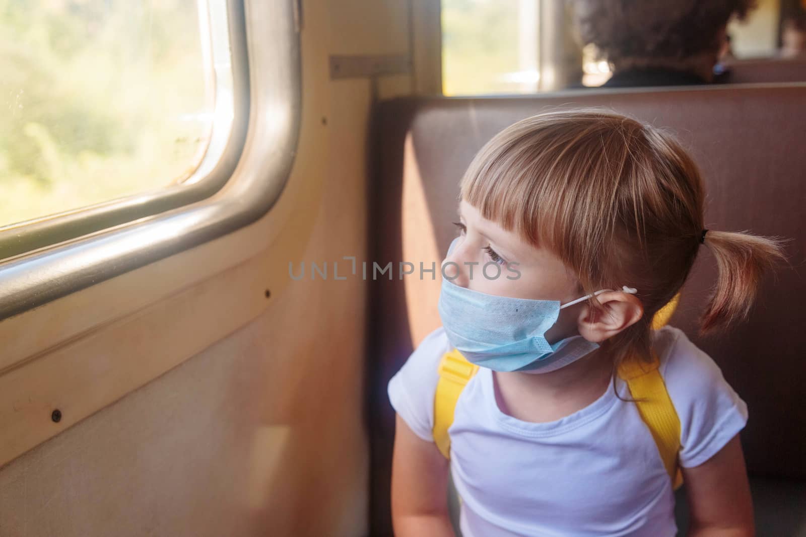 Little girl in a T-shirt and jeans and a medical mask is sitting in the train. by galinasharapova