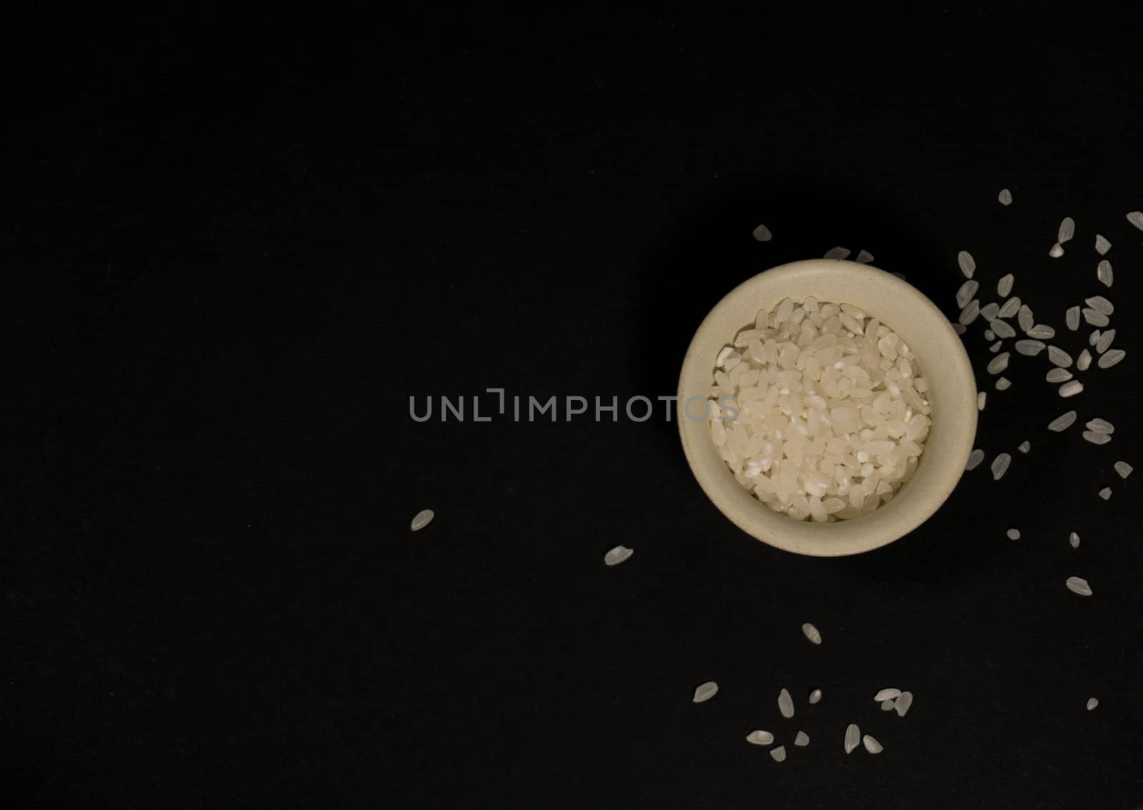 White uncooked rice in a beige bowl with scattered grains nearby on a black background