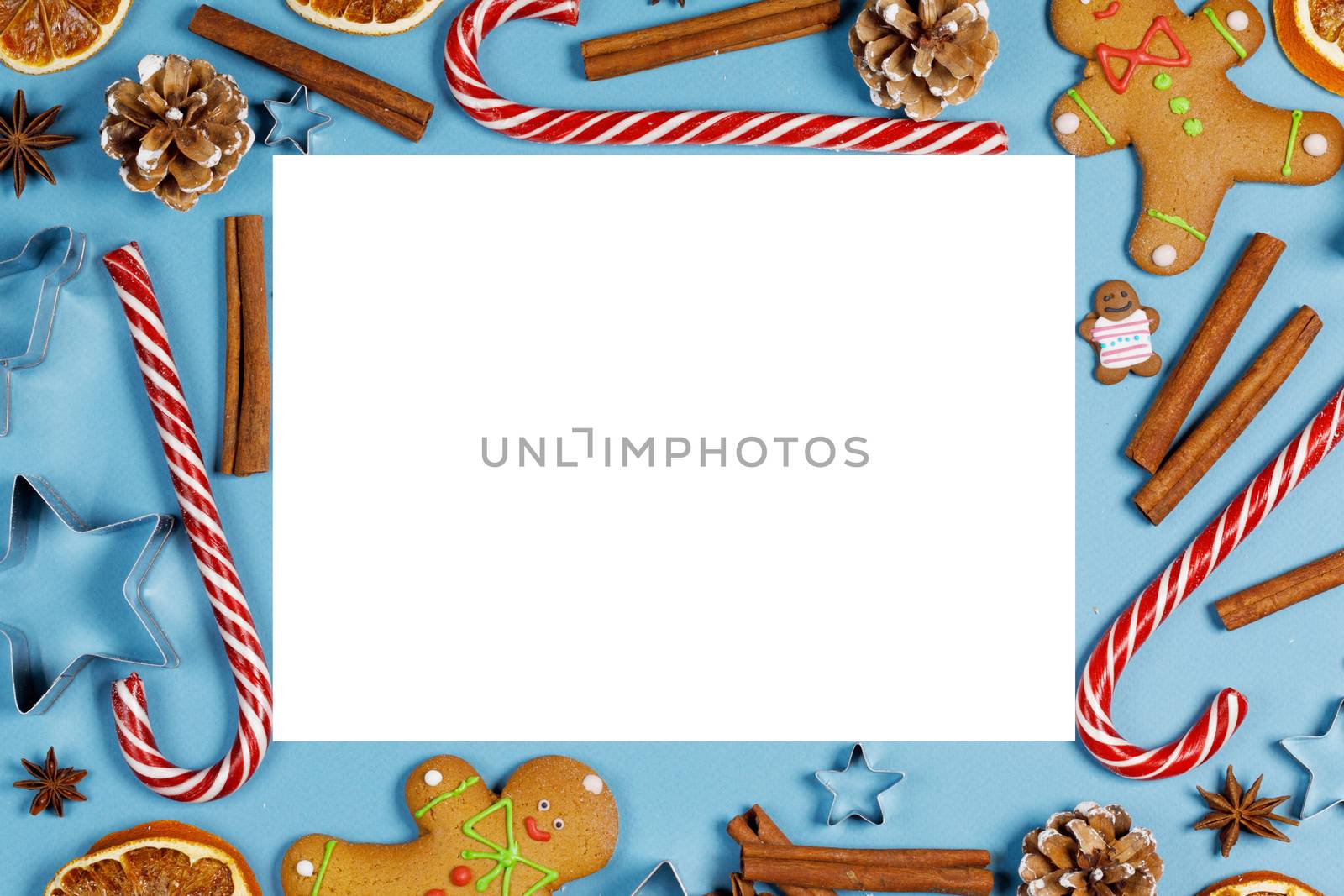 Frame of Christmas gingerbread cookies on blue background with copy space. Pattern of gingerbread men, candy canes, star mold, cinnamon, pine cone, orange. Top view, flat lay.