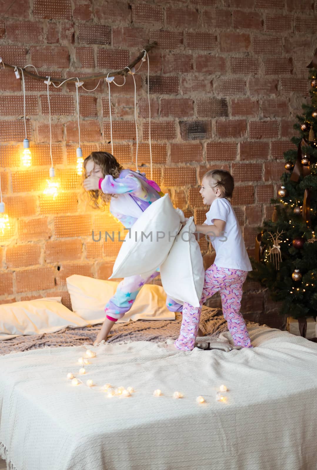.Little sisters in pajamas on the bed fight with pillows against the background of christmas garlands
