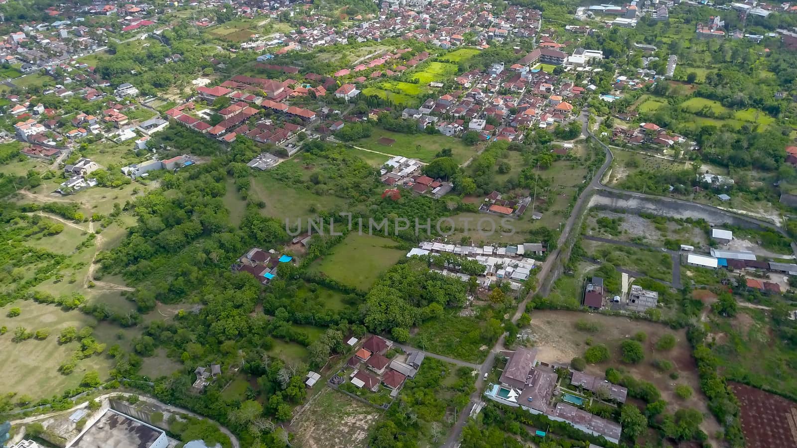 Many villas with brown-orange shingle roofs between tropical trees on the sky background in Ubud on Bali. Sun is shining onto them. Aerial photo.