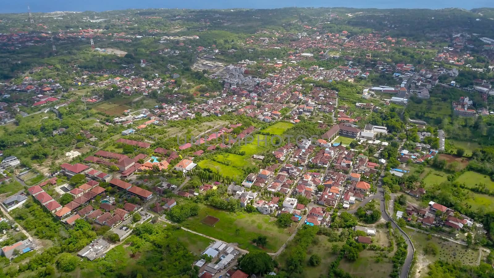 Many villas with brown-orange shingle roofs between tropical trees on the sky background in Ubud on Bali. Sun is shining onto them. Aerial photo by Sanatana2008