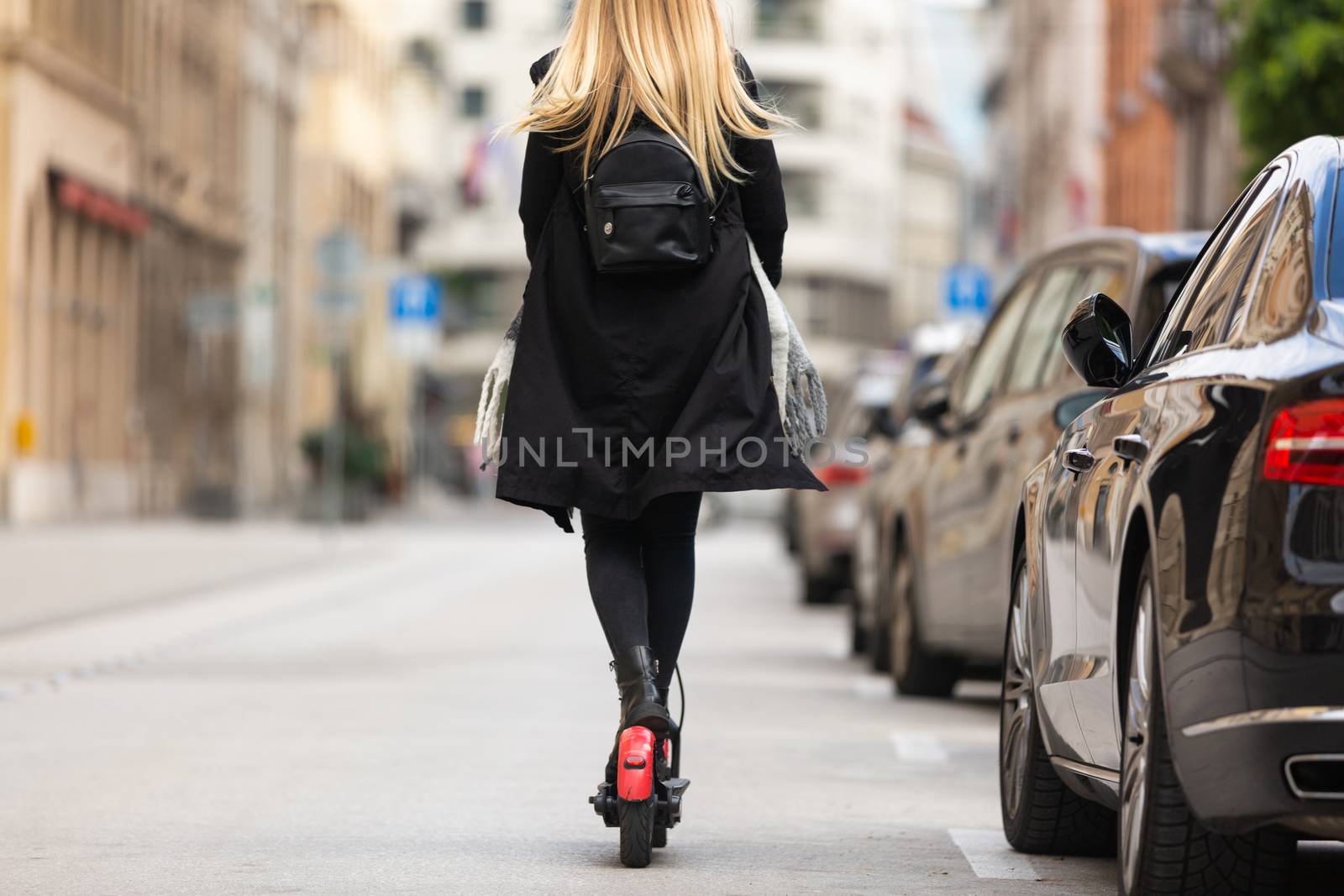 Rear view of girl riding public rental electric scooter in urban city environment at fall. New eco-friendly modern public city transport in Ljubljana.