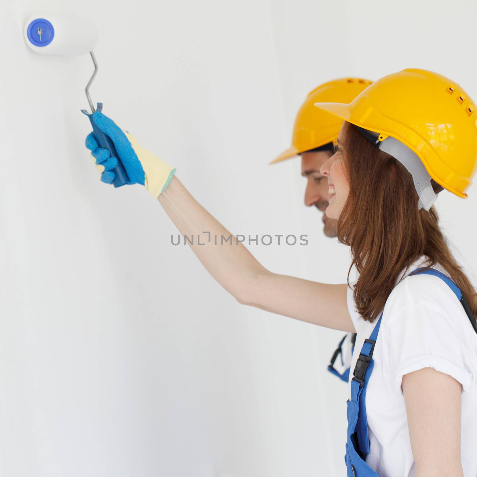 Two young workers in uniform of coverall and hardhat painting white wall