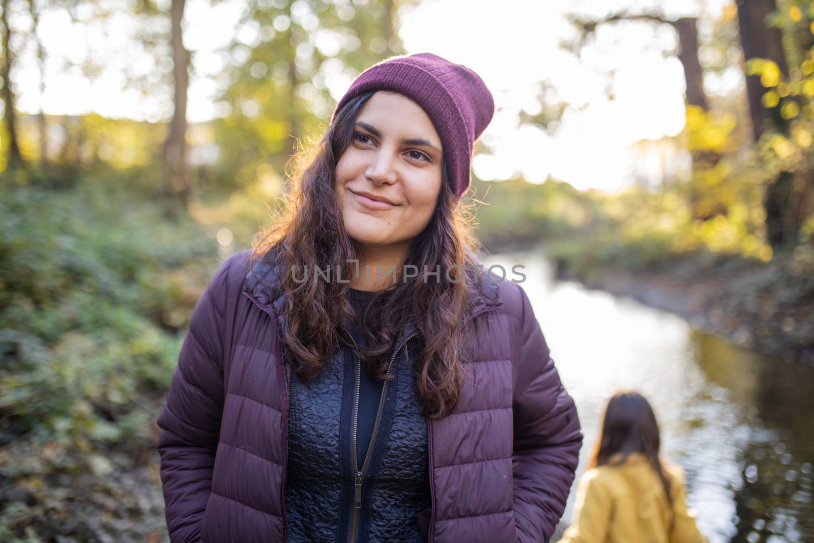 Happy brunette woman wearing knitted hat and standing next to a river in the forest. Portrait of woman in forest with river as background. Adventurous day in nature