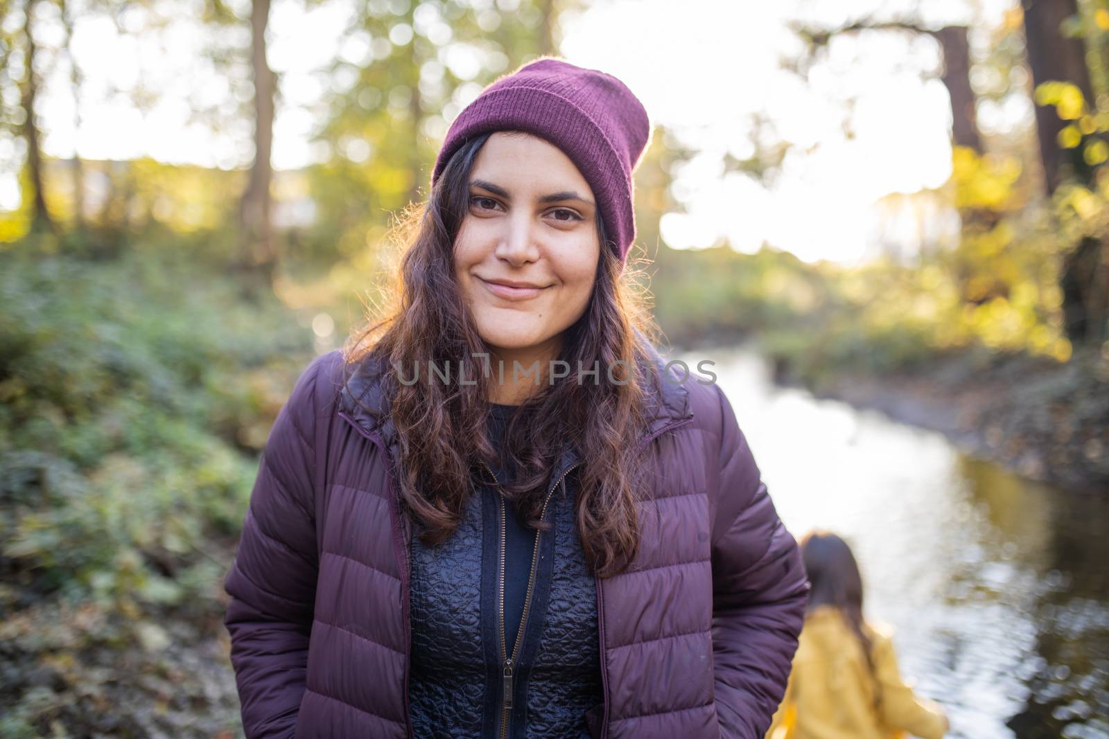 Happy brunette woman wearing knitted hat and standing next to a river in the forest. Portrait of woman in forest with river as background. Adventurous day in nature