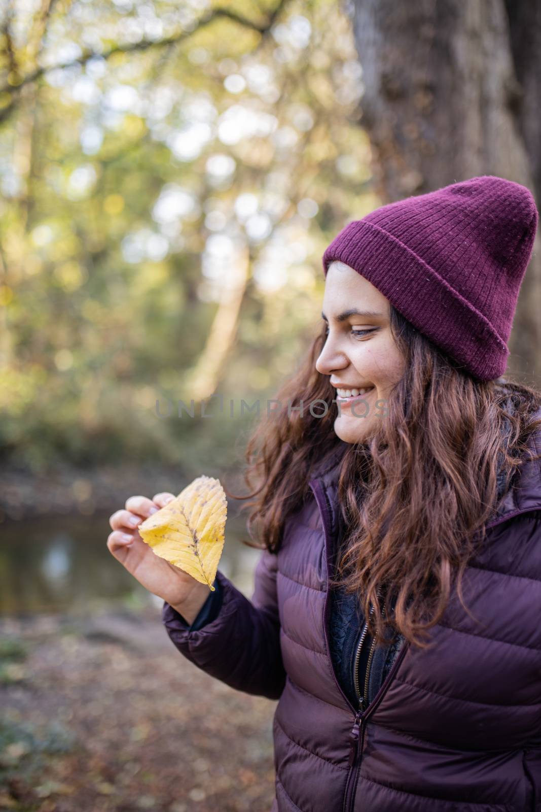 Happy woman smiling and holding an autumn leaf in the forest. Portrait of woman wearing knitted hat in forest. Adventurous day in nature