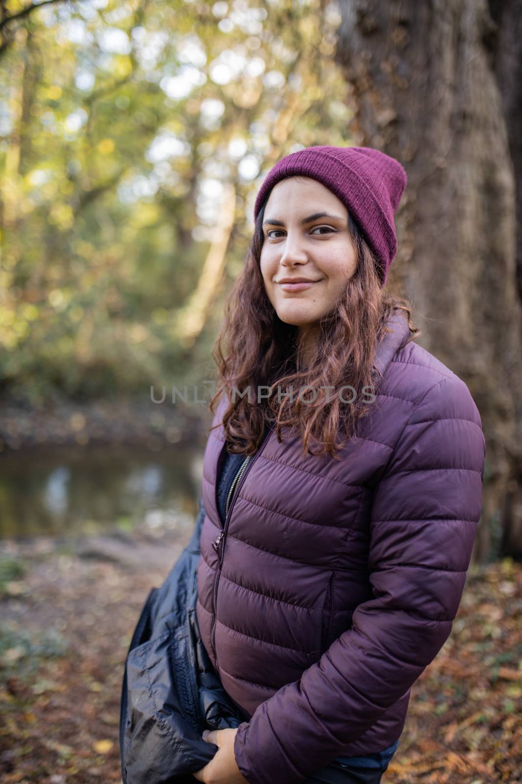 Happy woman with serene smile in the forest with a river as background. Portrait of woman wearing knitted hat in forest. Adventurous day in nature
