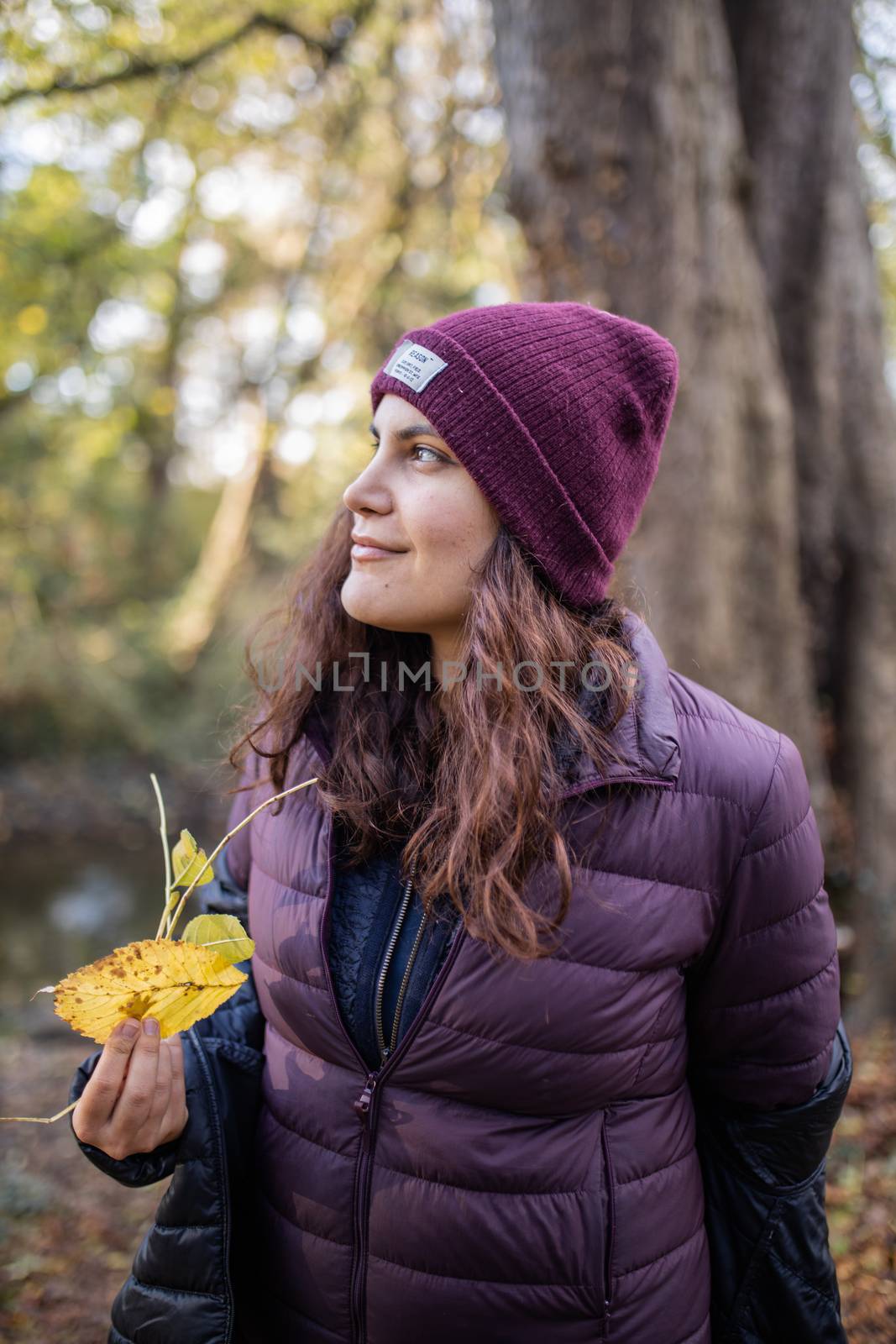 Smiling brunette woman holding a yellow leaf and looking away in the forest. Portrait of woman wearing knitted hat in forest. Adventurous day in nature