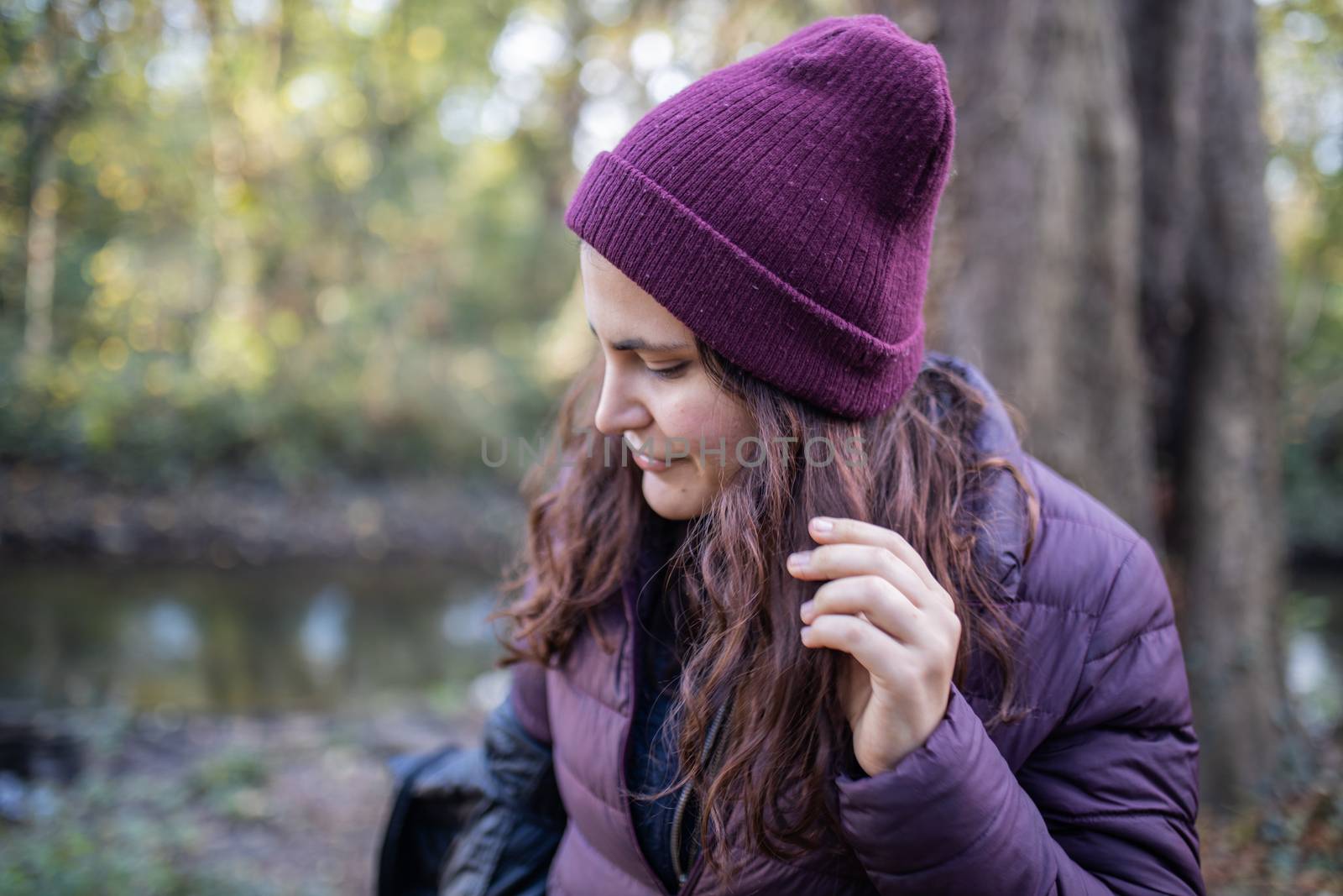 Smiling brunette woman looking away and wearing knitted hat next to a river in the forest. Portrait of woman in forest with river as background. Adventurous day in nature
