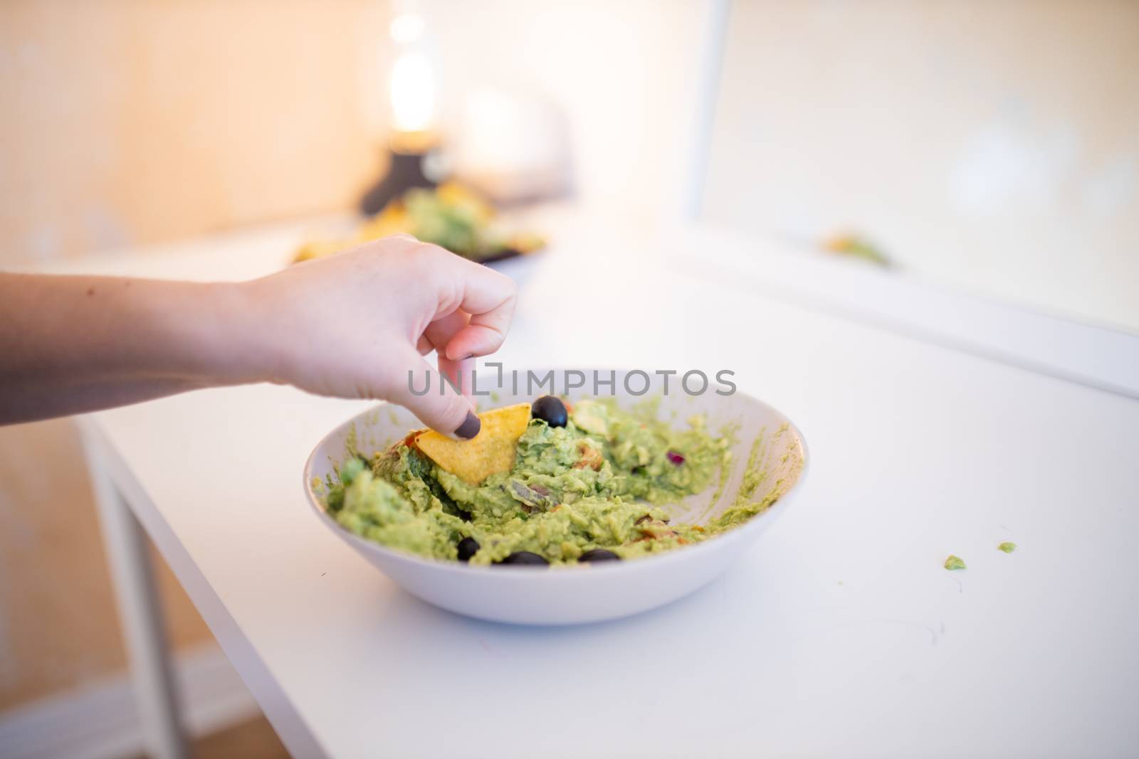 Female hand dipping nachos in guacamole sauce with olives on a white table. Bowl of traditional Mexican guacamole on a table. Spicy Mexican cuisine