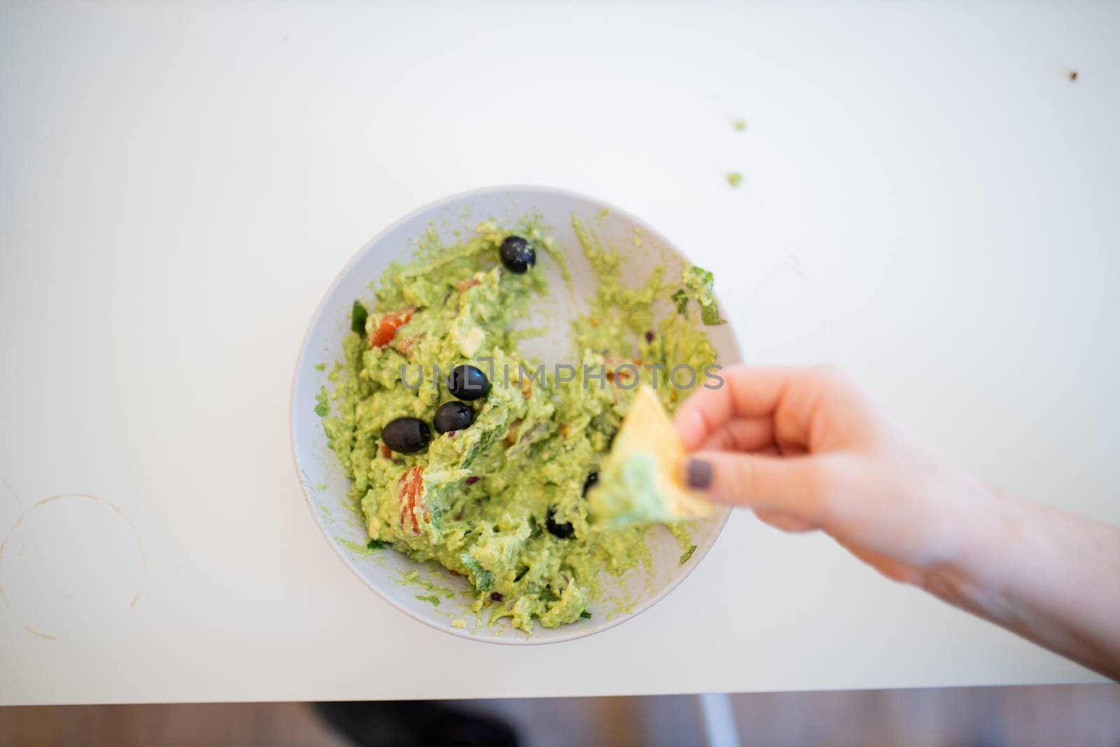 Female hand dipping nachos in guacamole sauce with olives on a white table. Traditional Mexican guacamole on a table from above. Mexican cuisine