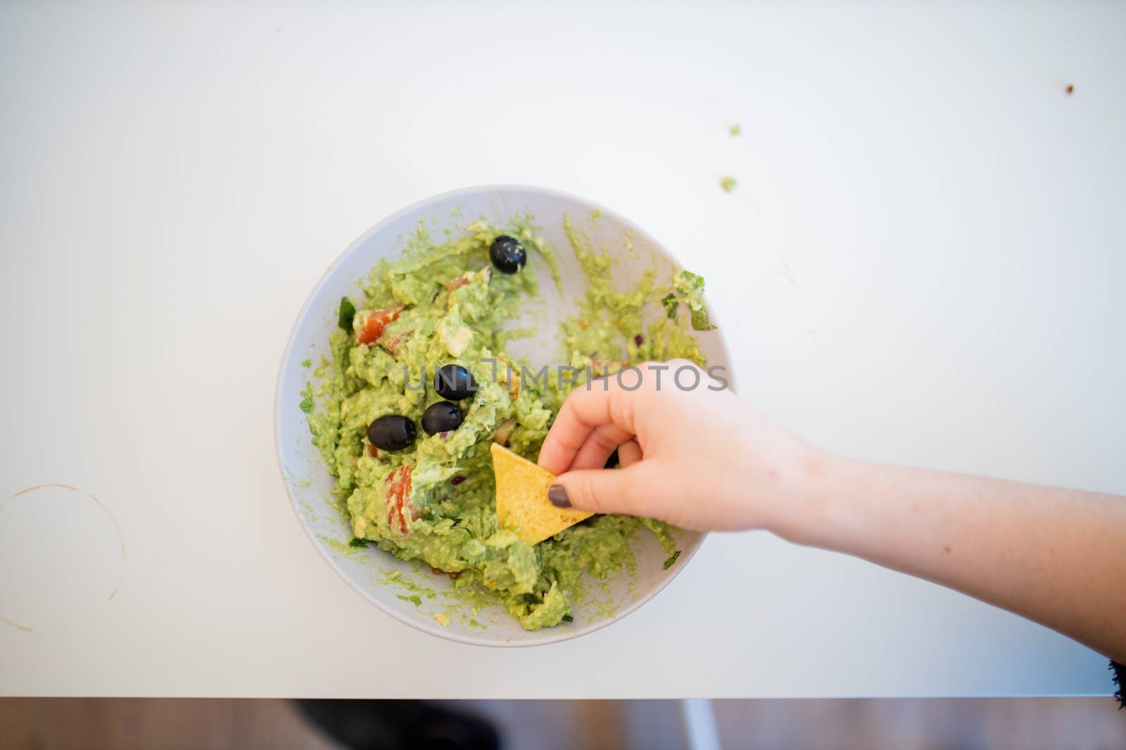 Female hand dipping nachos in guacamole sauce with olives on a white table. Traditional Mexican guacamole on a table from above. Spicy mexican cuisine