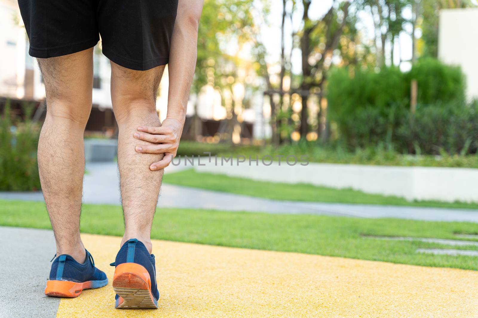 Male runner athlete leg injury and pain. Hands grab painful leg while running in the park. by mikesaran