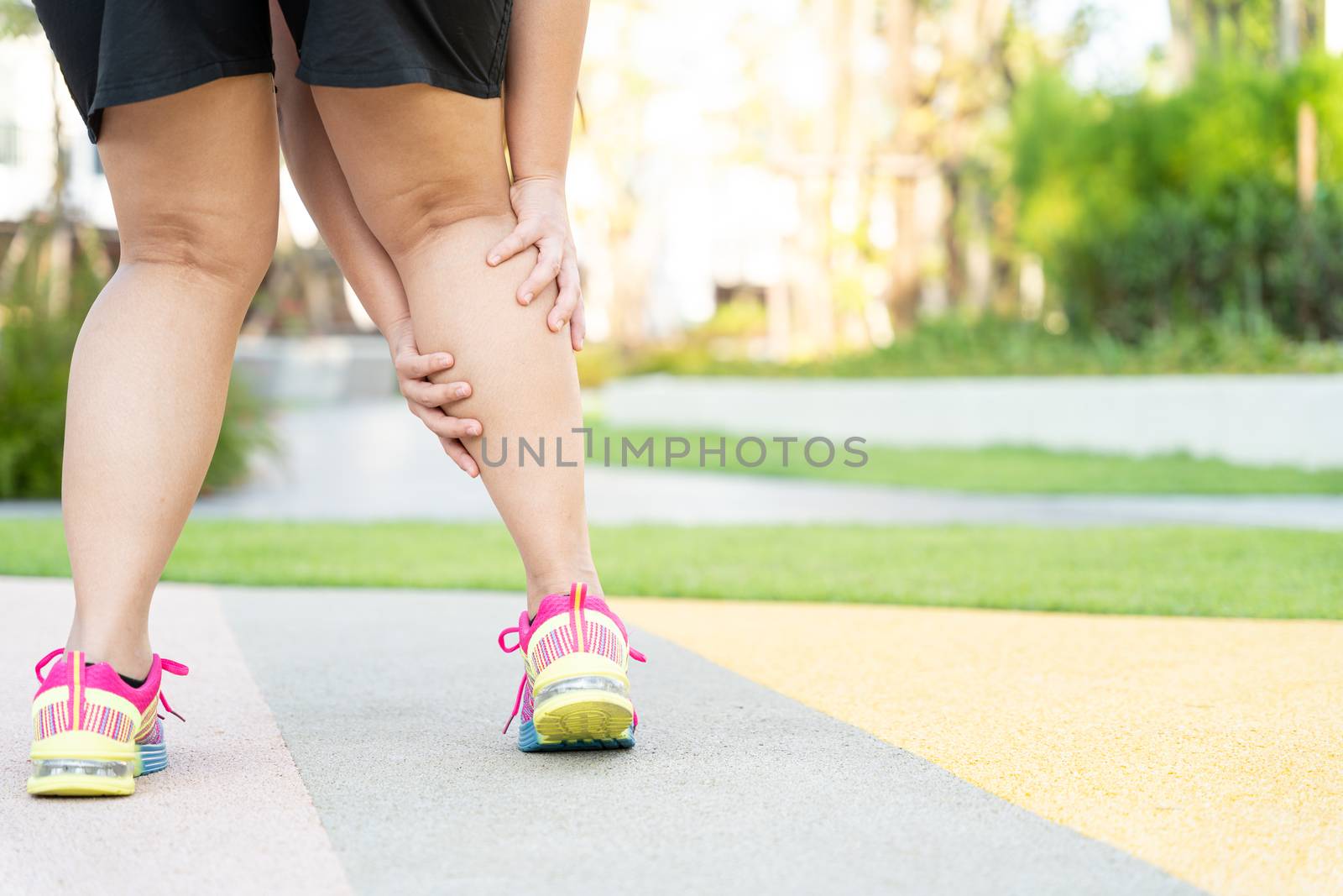 Female fatty runner athlete leg injury and pain. Hands grab painful leg while running in the park.