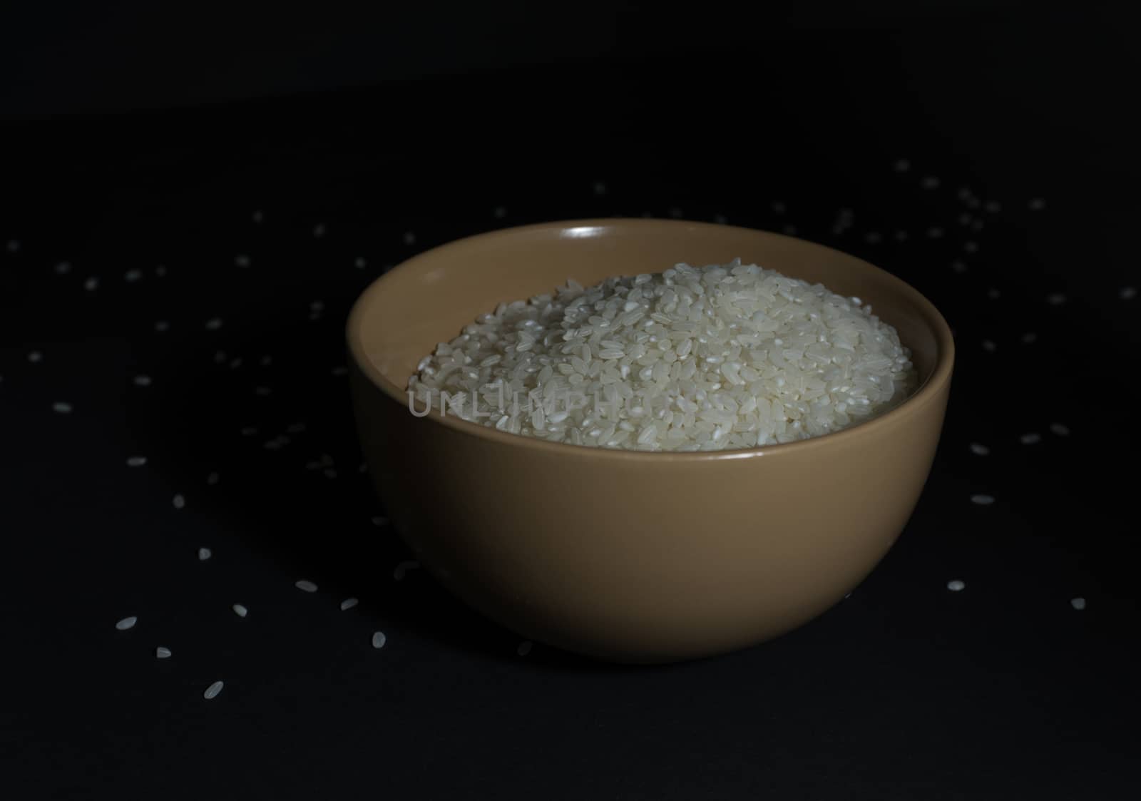 White uncooked rice in a beige bowl with scattered grains nearby on a black background