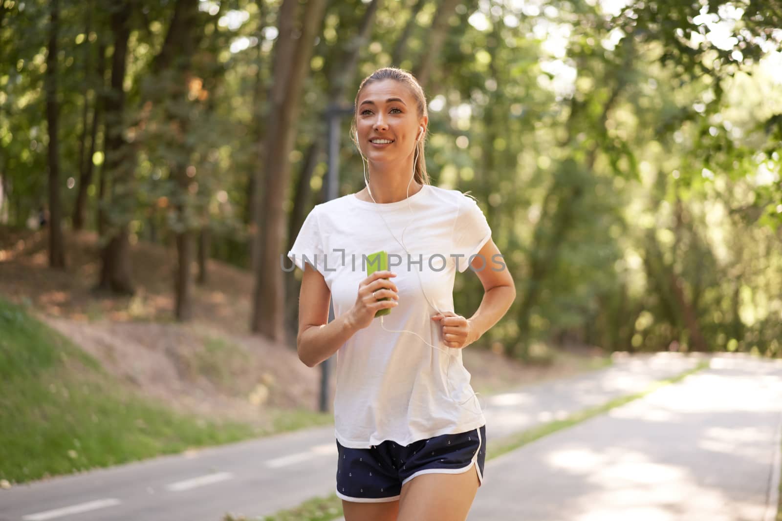 Woman listen music running on asphalt track summer park Caucasian female earphones jogging sunny morning Runner training hold smartphone in hand Healthy lifestyle positive concept