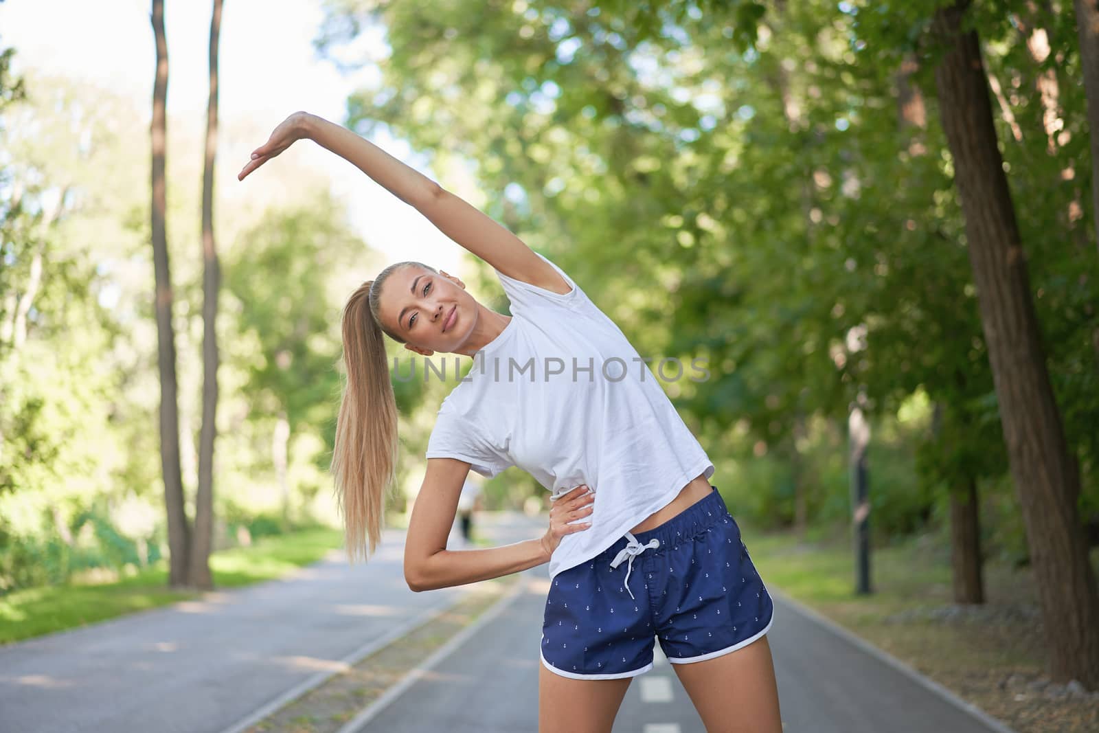Woman runner stretching arms before exercising summer park morning Middle age athletic female warming up body before running Caucasian person warm up jogging Dressed white shirt shorts running track