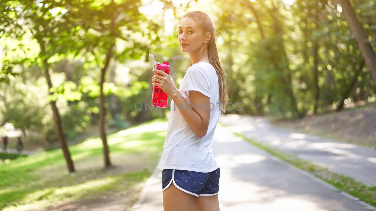 Woman drink water red bottle after morning workout. Young athletic female rear view walk asphalt running track park after jogging Healthy lifestyle concept
