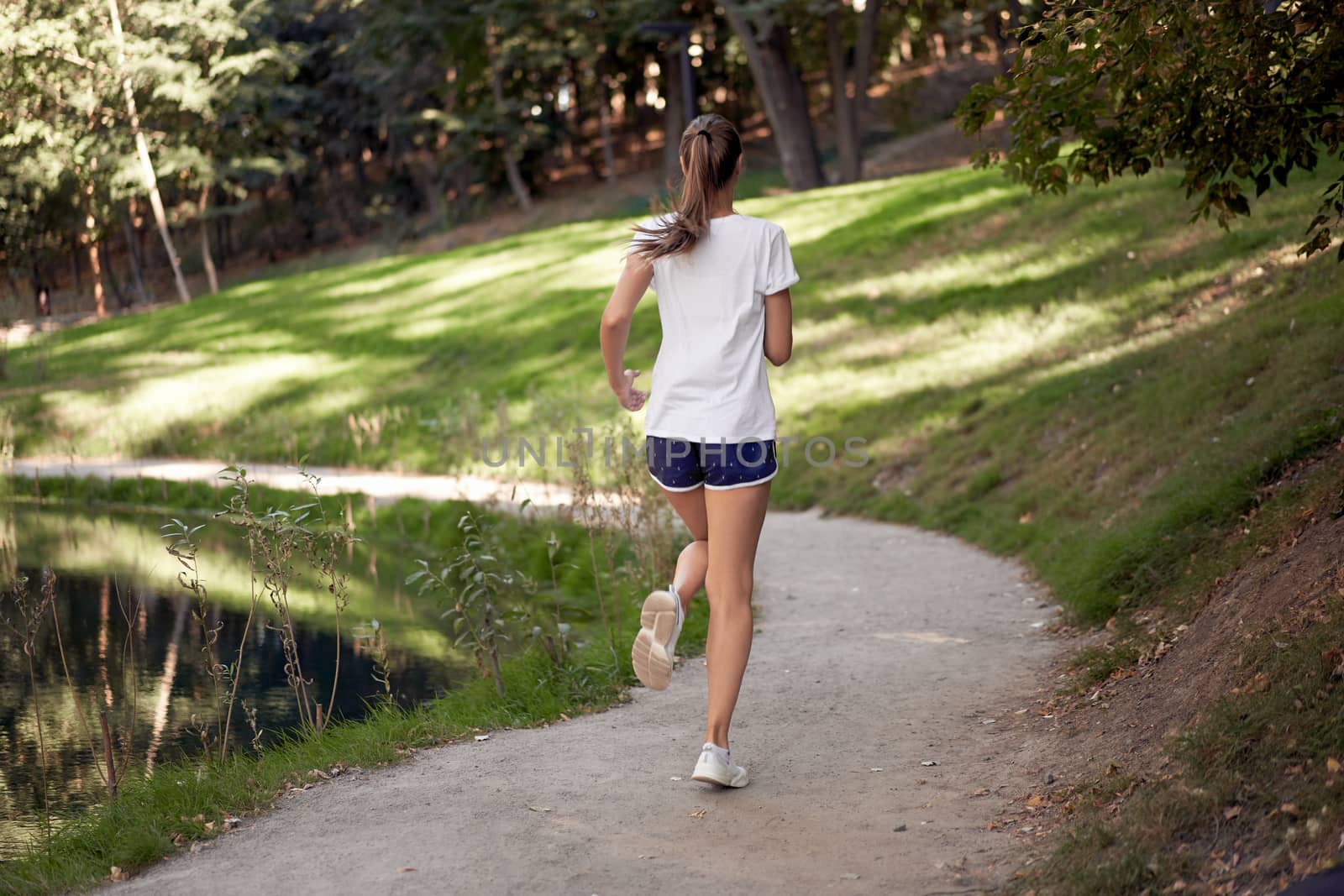 Woman running road summer park near lake Active sporty caucasian female morning workout Healthy lifestyle concept. Athletic person sportswear exercises Dressed white t-shirt full length Rear back view