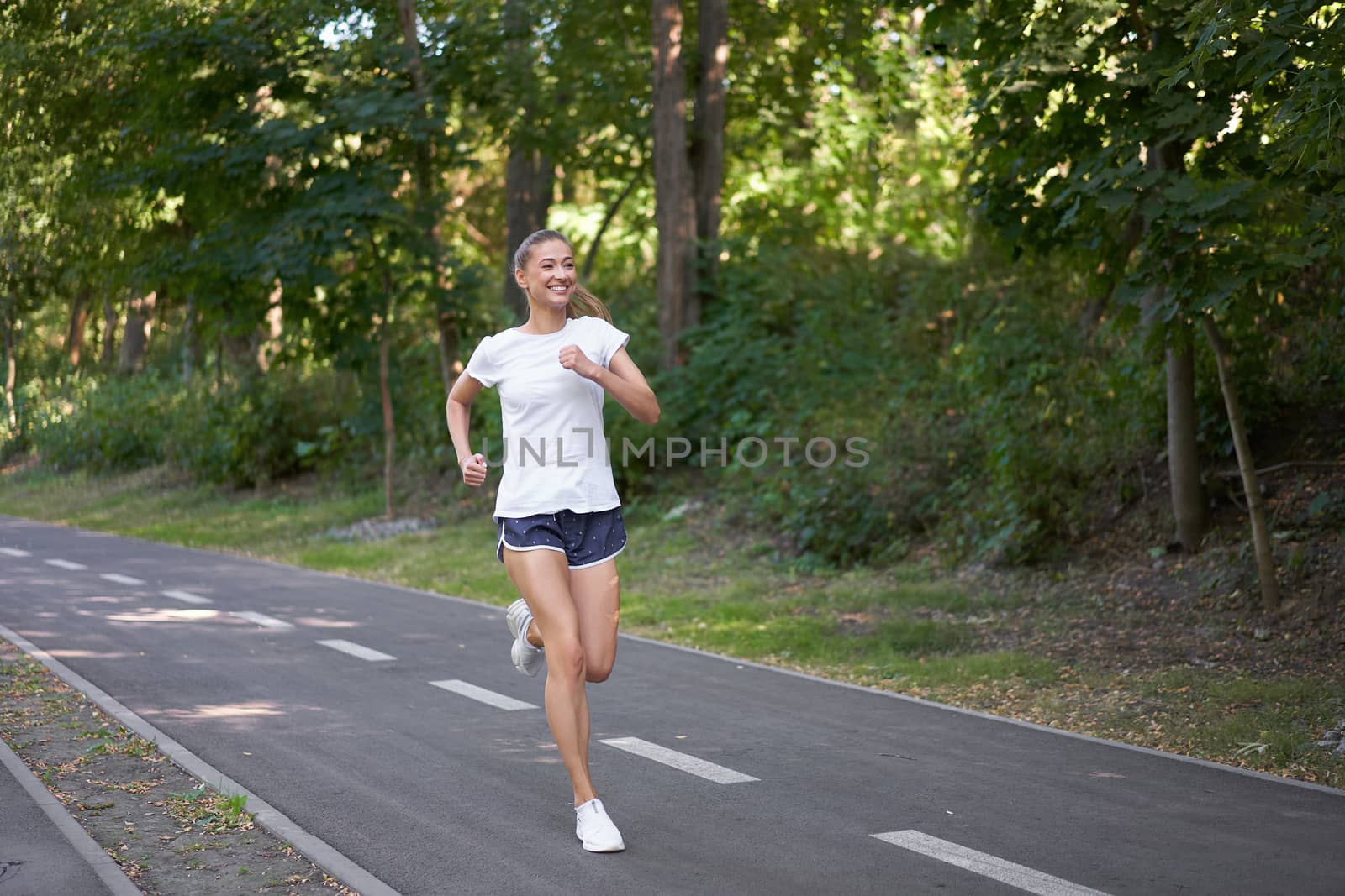 Woman running asphalt road summer park Active sporty caucasian female morning workout Healthy lifestyle concept. Athletic person sportswear exercises Dressed white t-shirt full length