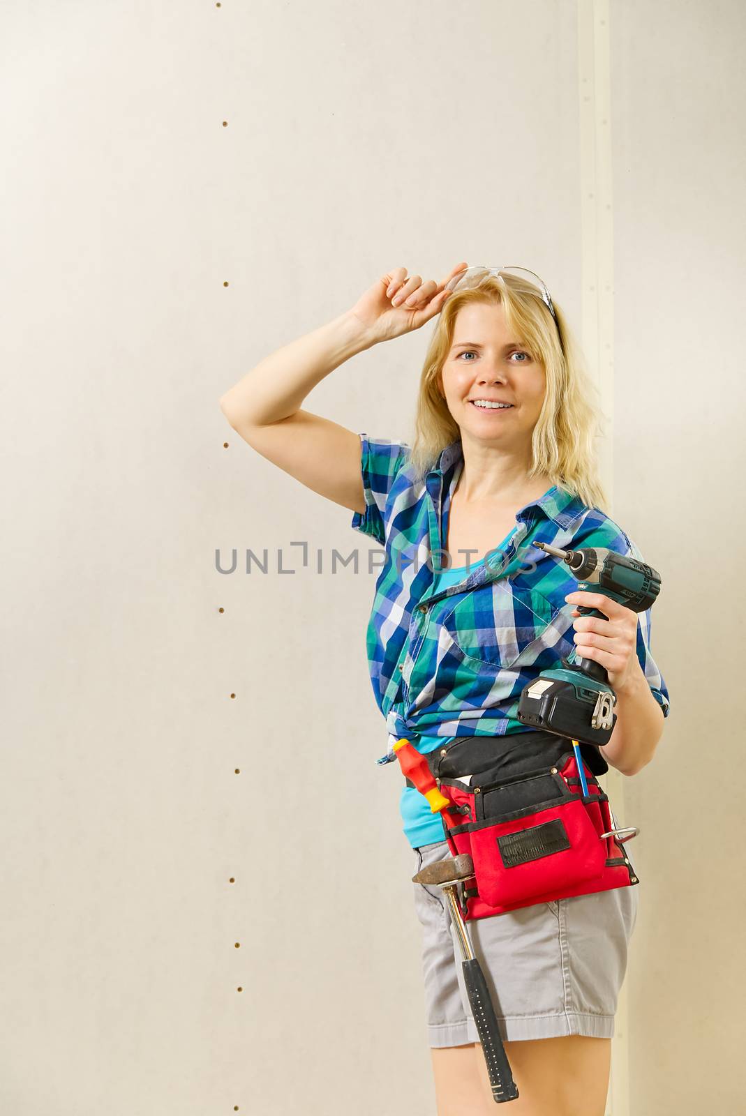 Blond woman wearing a DIY tool belt full of a variety of tools on a unpainted plasterboard wall background. Construction woman concept. by PhotoTime