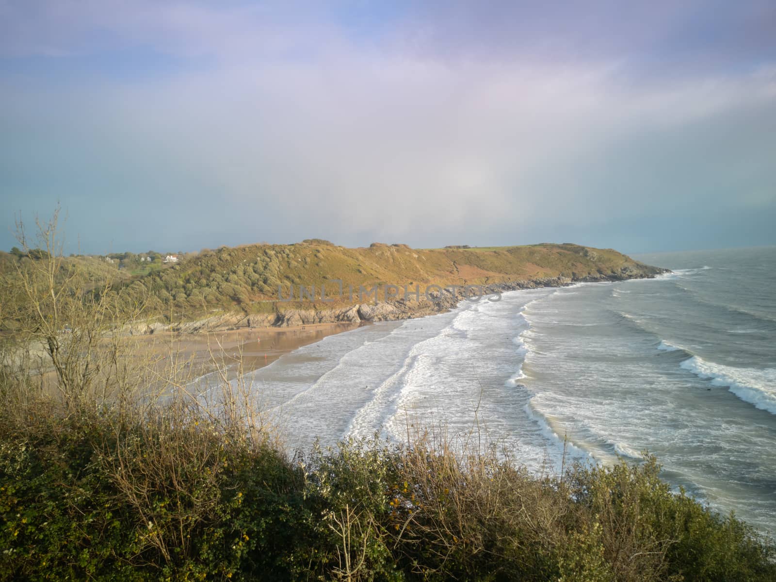 Approaching high-tide at Caswell Bay Beach in Wales, UK. Stunning view of crashing waves onto the sandy shores from the Gower Coastal Path towards sunset.