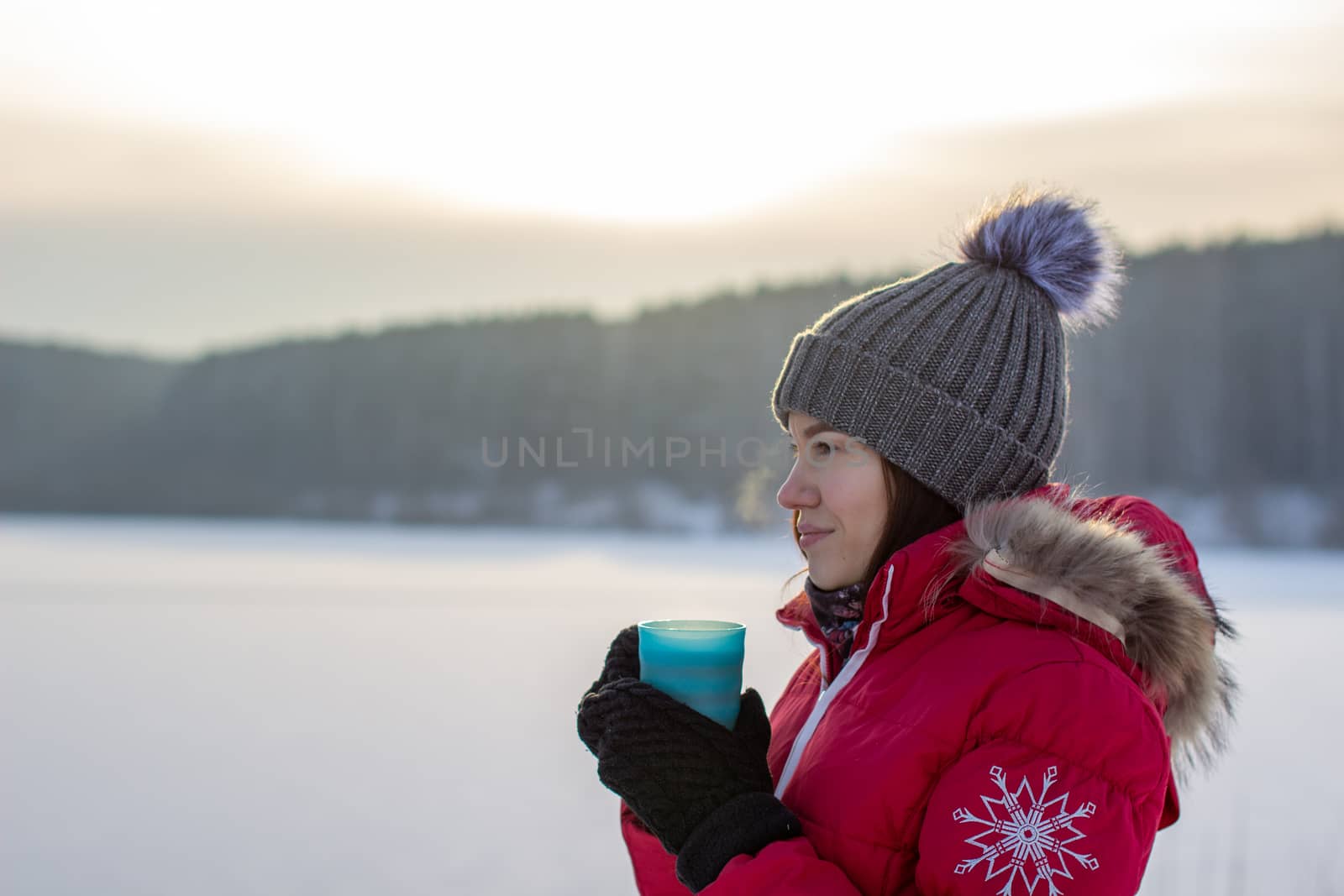Beautiful girl in a winter jacket with a mug in winter in nature. A girl in a hat and a red warm jacket. He warms his hands and drinks hot tea or a drink 