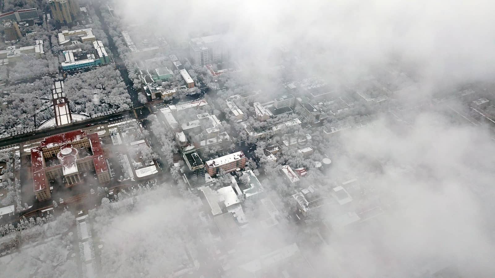 Top view through the clouds on the city of Almaty. Winter atmosphere, white trees, bushes, roofs of houses. Flying over the city. White clouds and light fog. Traffic of cars on roads. Through clouds.