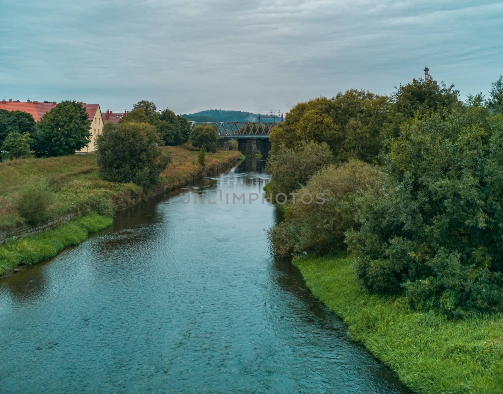 Cloudy Landscape of small river between bushes and trees with train bridge over it by Wierzchu