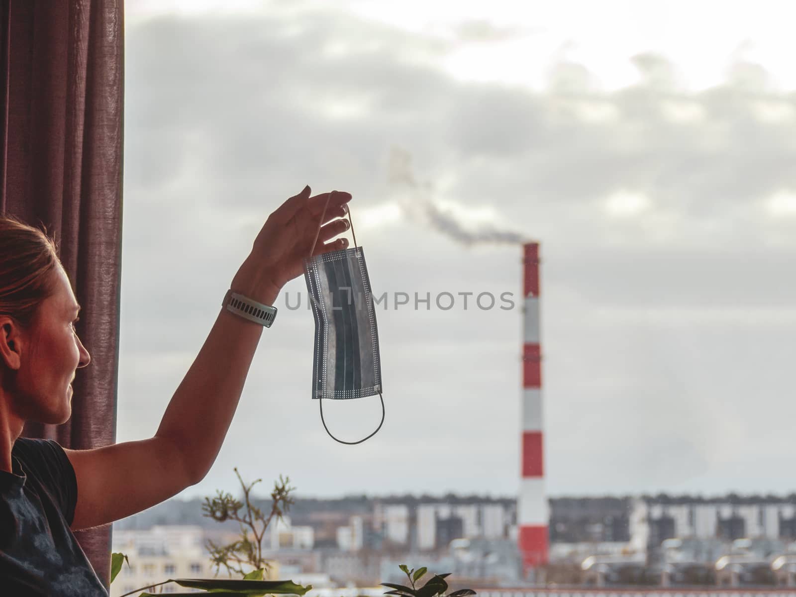 Young Woman Hold In Hands Medical Mask . Smoking factory big tall pipe. cloudy sunset sky on background. Rainy day. Concept of ecology problems. Filtered image. copy space. Toned image