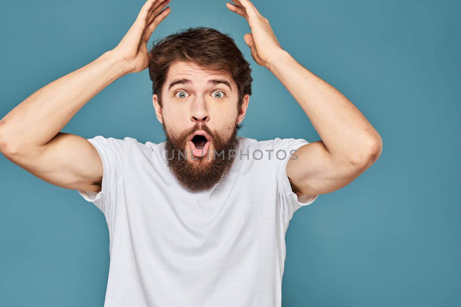 A bearded man in a white T-shirt gestures with his hands emotions blue background. High quality photo