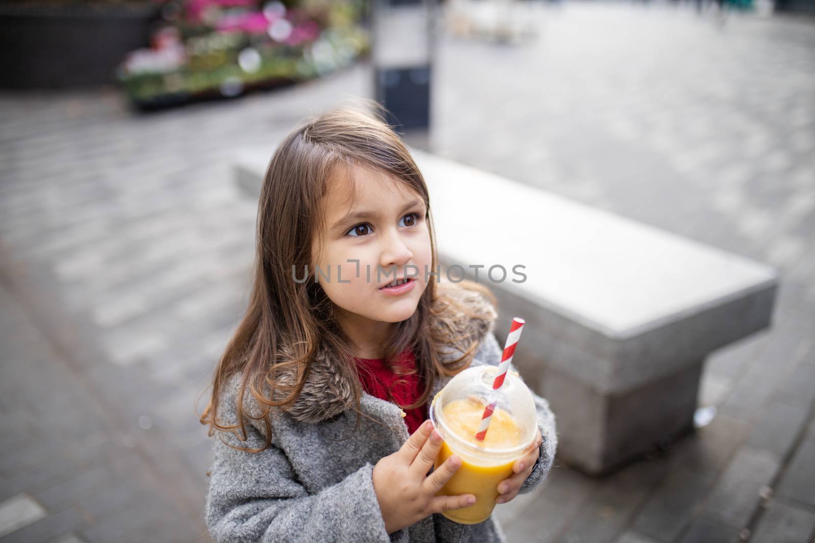 Adorable brunette little girl in the street holding yellow smoothie with striped straw. Cute young child holding a cold fruit beverage. Refreshing drinks for kids