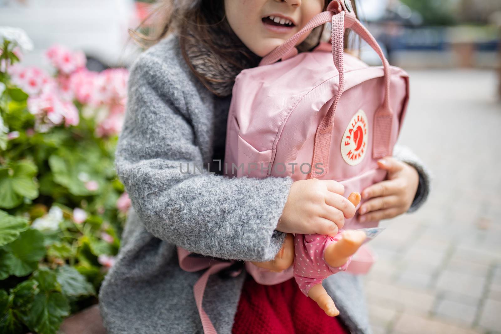 London, UK - November 4, 2020: Happy little girl with flowers behind her hugging pink Fjallraven backpack and small doll. Young child holding pink rucksack and doll. School supplies for children