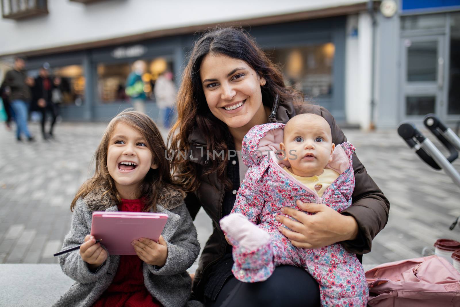Happy smiling mother sitting next to happy little daughter and holding her cute baby. Joyful mother hugging daughters outside. Young family fun day outdoors