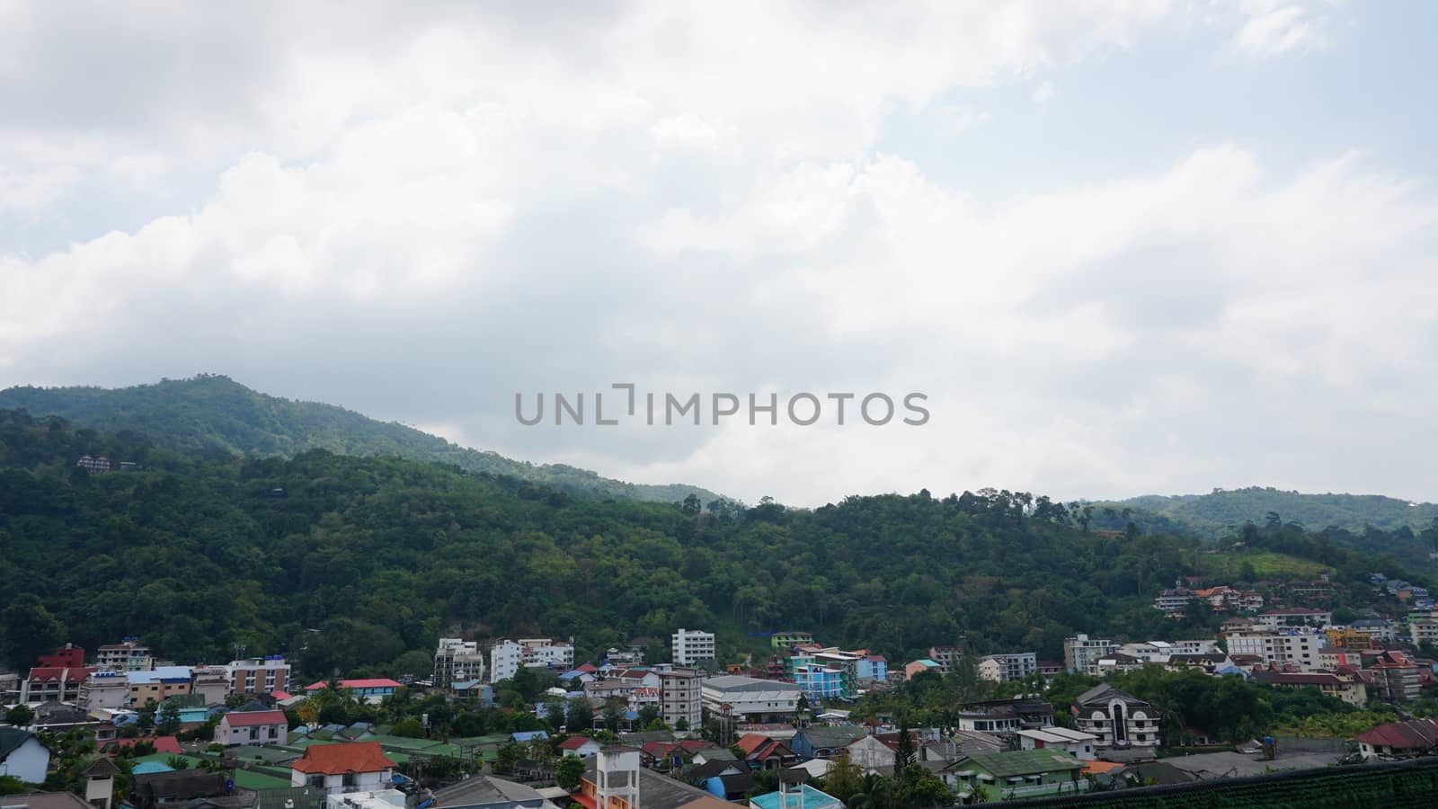 Drone view of the city of Patong, Phuket island. Houses of different heights stand on the beach. Green hills of the island. On the roof of houses pools, people swim. On the road go scooters and cars.