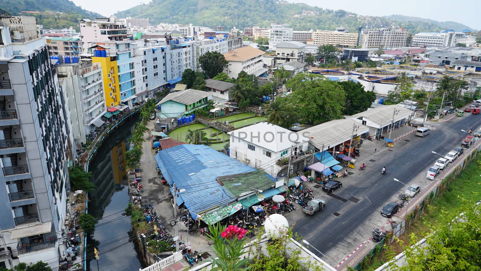Drone view of the city of Patong, Phuket island. Houses of different heights stand on the beach. Green hills of the island. On the roof of houses pools, people swim. On the road go scooters and cars.