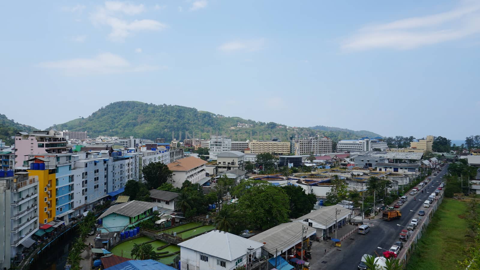 Drone view of the city of Patong, Phuket island. Houses of different heights stand on the beach. Green hills of the island. On the roof of houses pools, people swim. On the road go scooters and cars.