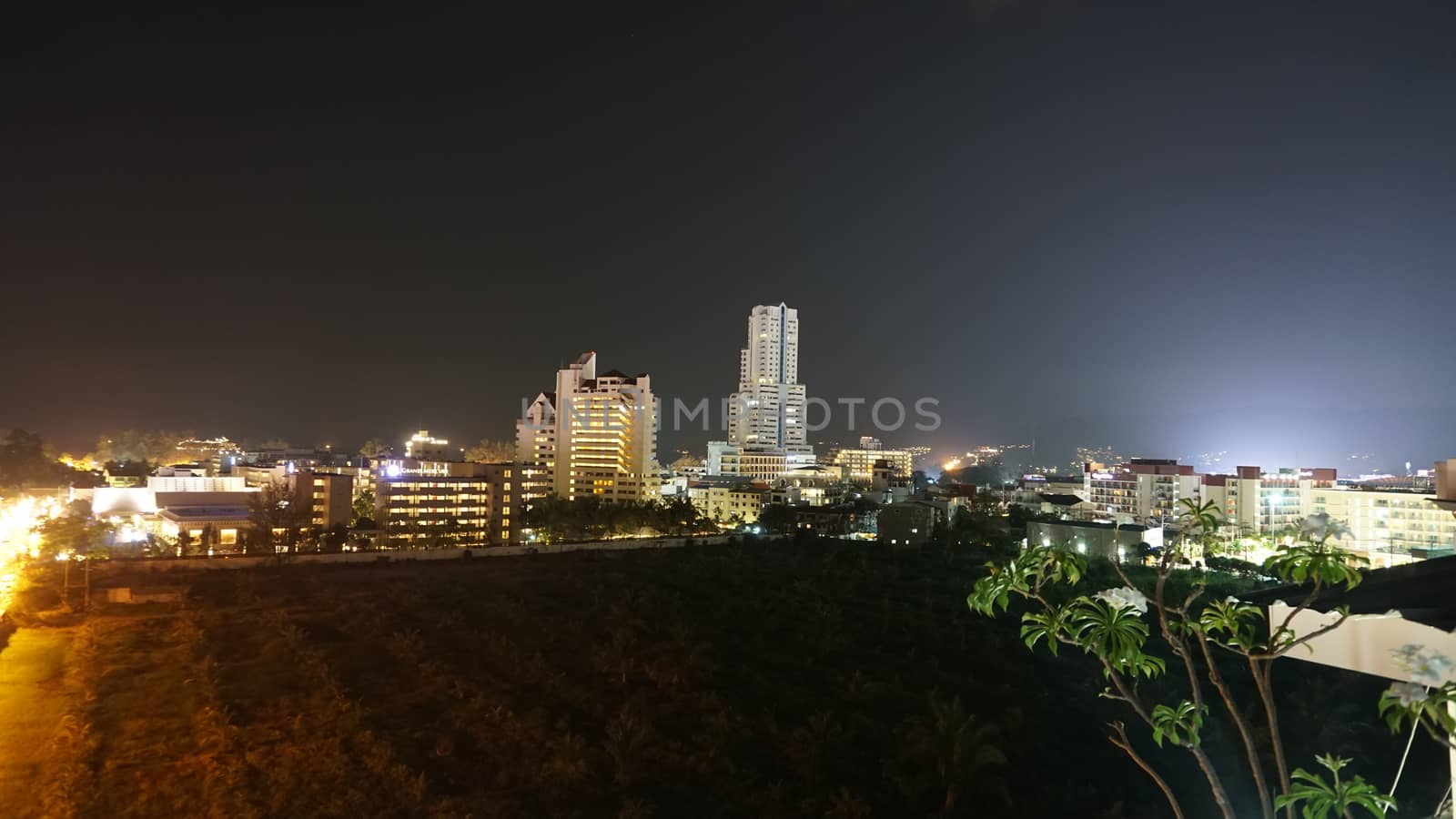 Night city of Patong, Phuket island. Houses of different heights stand on the beach. Street lights are shining brightly. On the road go scooters and cars. Tropical climates, green palm trees.