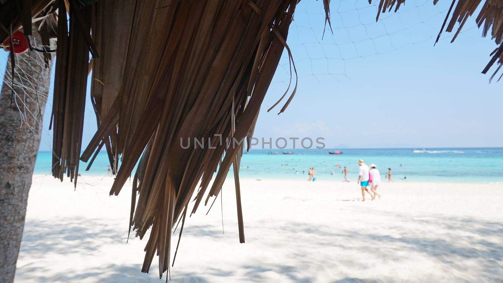 Palm leaves on the roof of the gazebo. Beach with white sand and blue water. People are actively spending time. Blue sky with clouds. The trunk of the palm tree. Bamboo Island, Thailand.