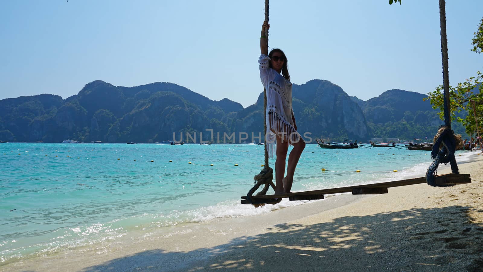 Beautiful girl in a white Cape and a black bathing suit on a swing. Swing on the beach. View of the island, beach with sand, blue water and green leaves of trees. Visible hills of the island. Thailand