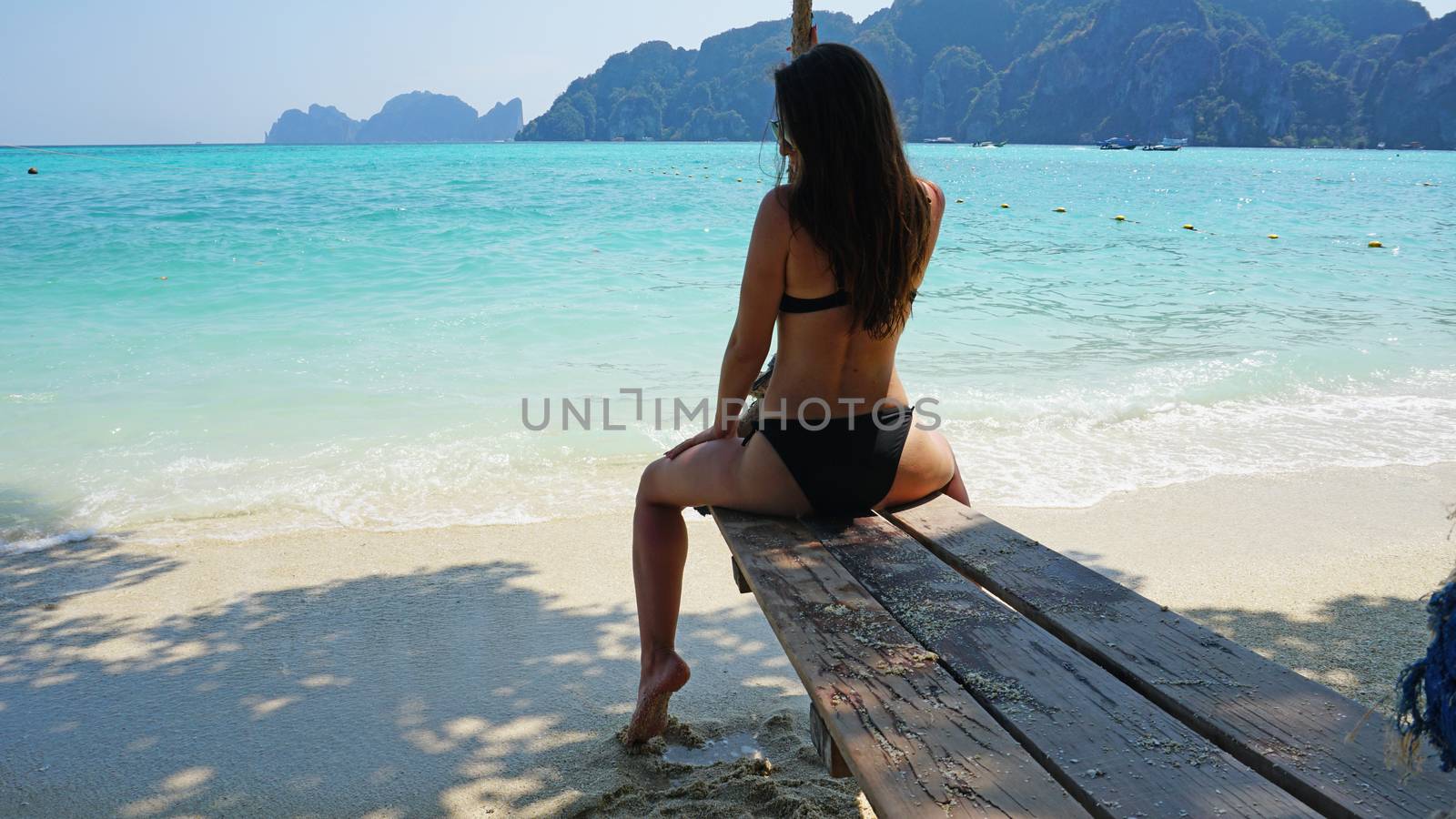 Girl in a swimsuit and glasses sitting on a swing. Swing on the beach. Black swimsuit, long hair. White sand and blue water. View of the blue sky and green Islands. Long rope. Thailand